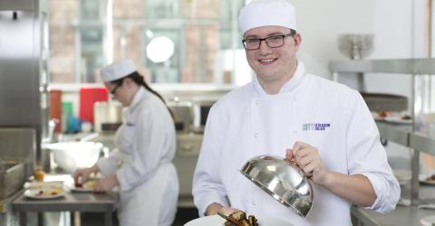 Two students wearing chefs whites in a professional kitchen.  One student in the background is cutting up food and one is holding a plate with food on it.